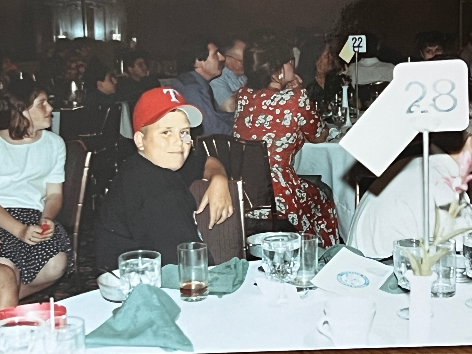 A quiet look across the room at a family celebration — cap on, smile half-hidden, taking it all in