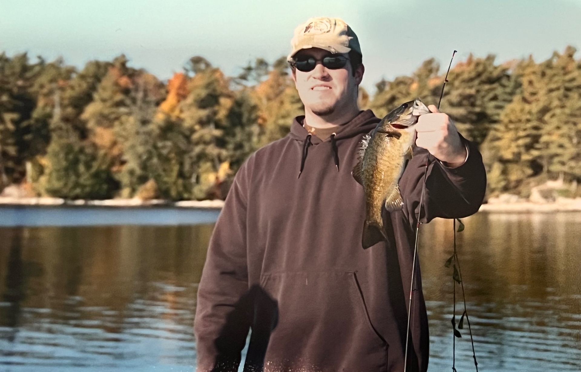 A proud smallmouth held high — autumn pines mirrored in the lake behind him