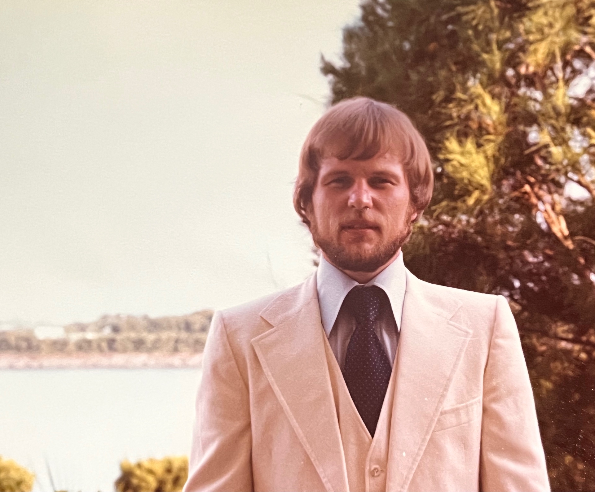 Cream three-piece suit, waterfront behind him — Richard just after the wedding, dressed sharp and stepping into his new life with Yvonne