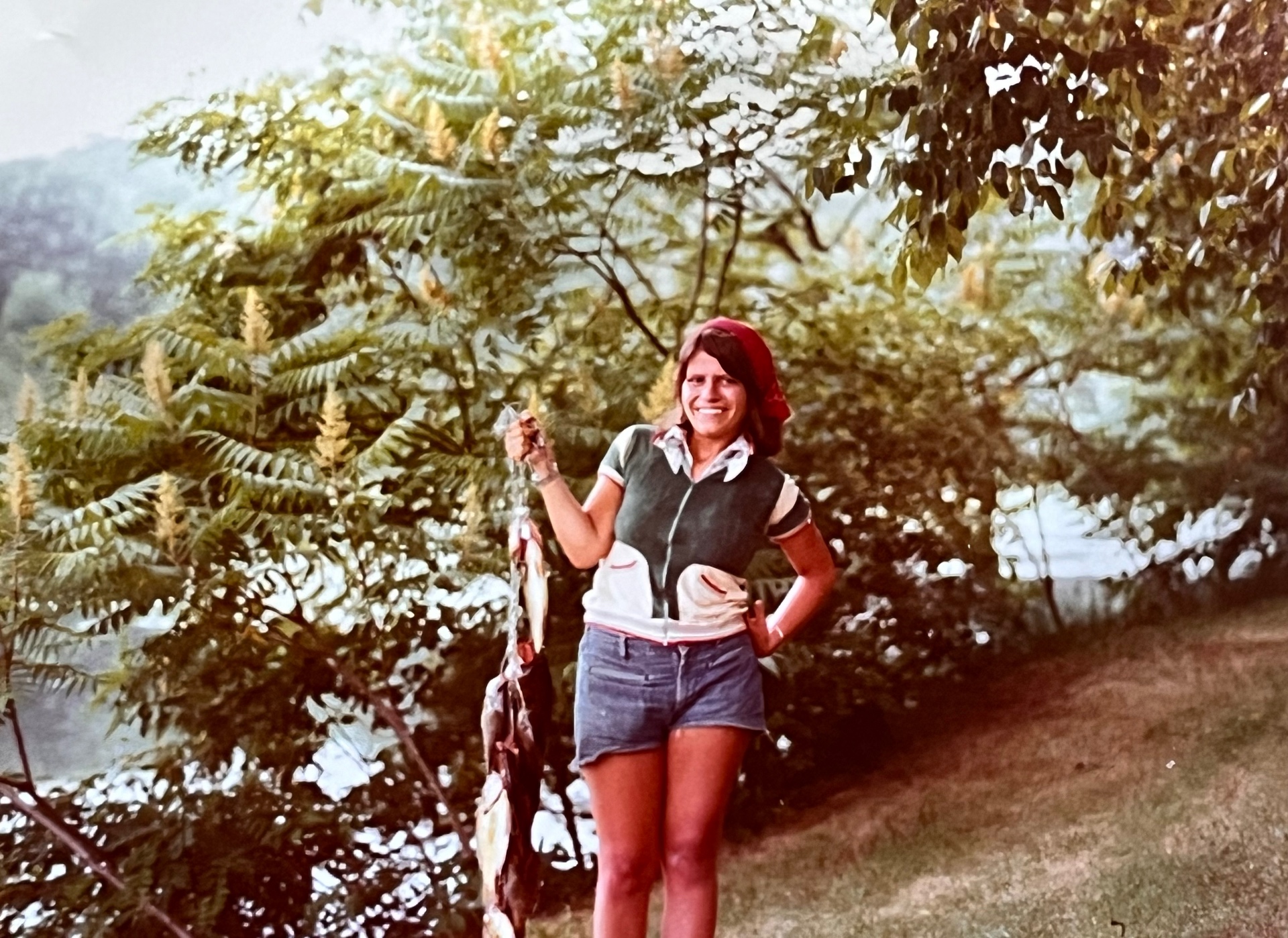 Yvonne on the riverbank with a full stringer — red bandana, denim shorts, biggest smile in the world. She fished right alongside him