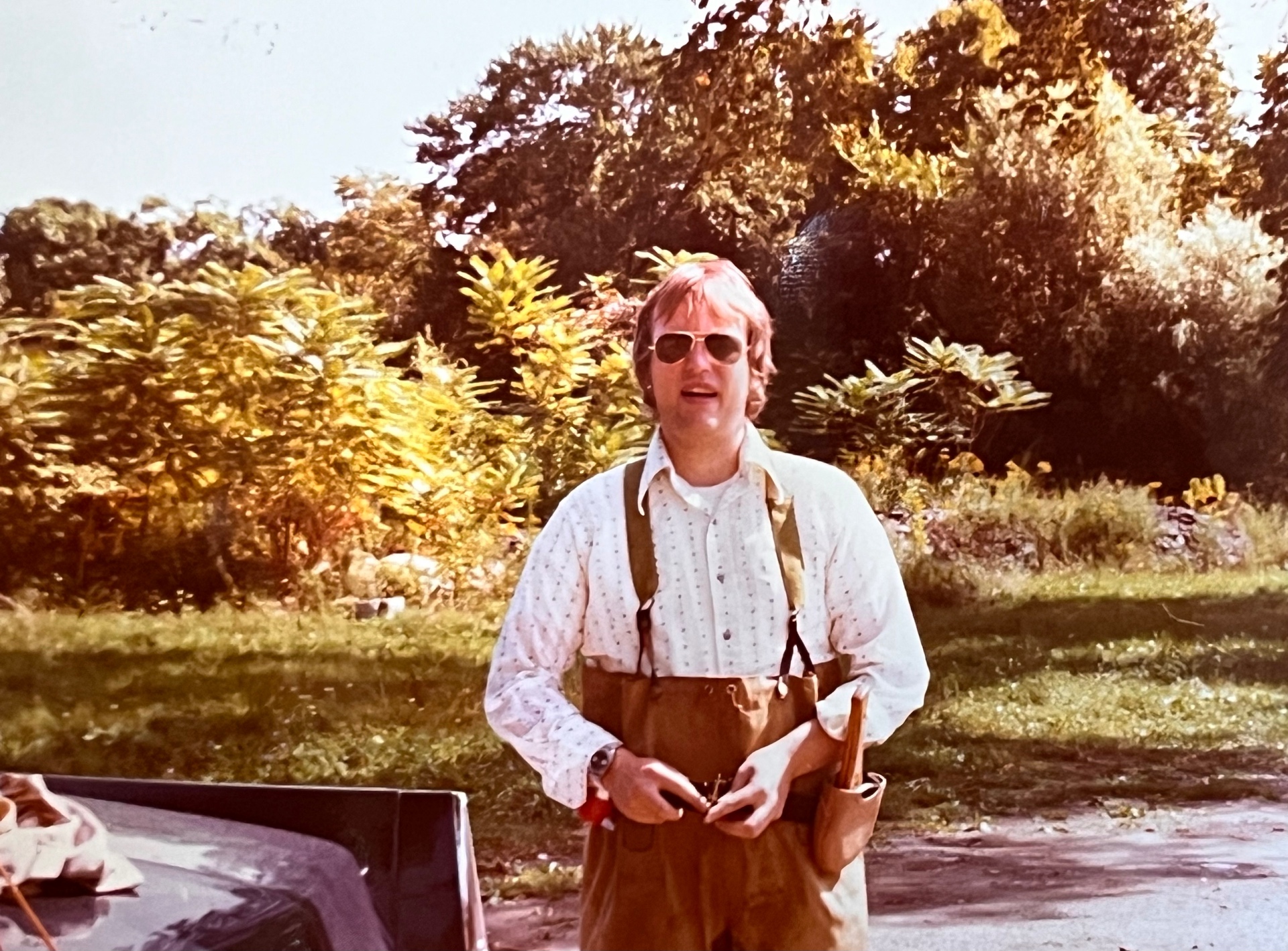Aviators, suspenders, waders rolled down — Richard back from the stream, gear still on, story ready to tell