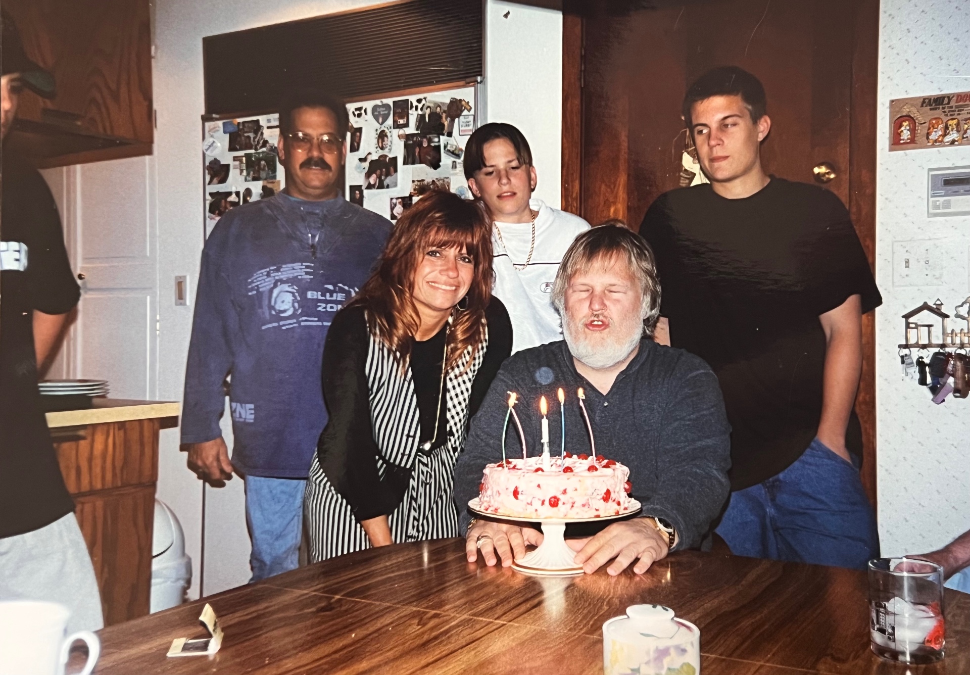 Candles glowing, the family gathered close around Richard — cherry cake in hand, everyone he loved in frame